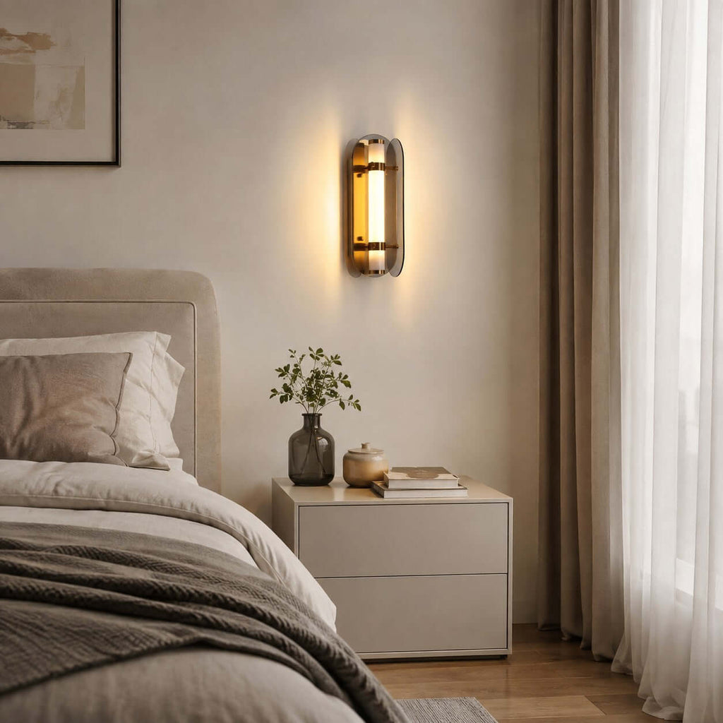 Clérachic wall light glowing above a pale bedside cabinet, with a small plant in a vase and a cream bed with sheer curtains.