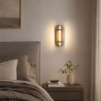 Clérachic wall light with smoked glass wings lit beside a grey upholstered headboard and a bedside table with a vase.