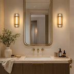 A pair of lit Clérachic wall lights flank a tall brass-framed mirror above a stone bathroom vanity with brass taps.