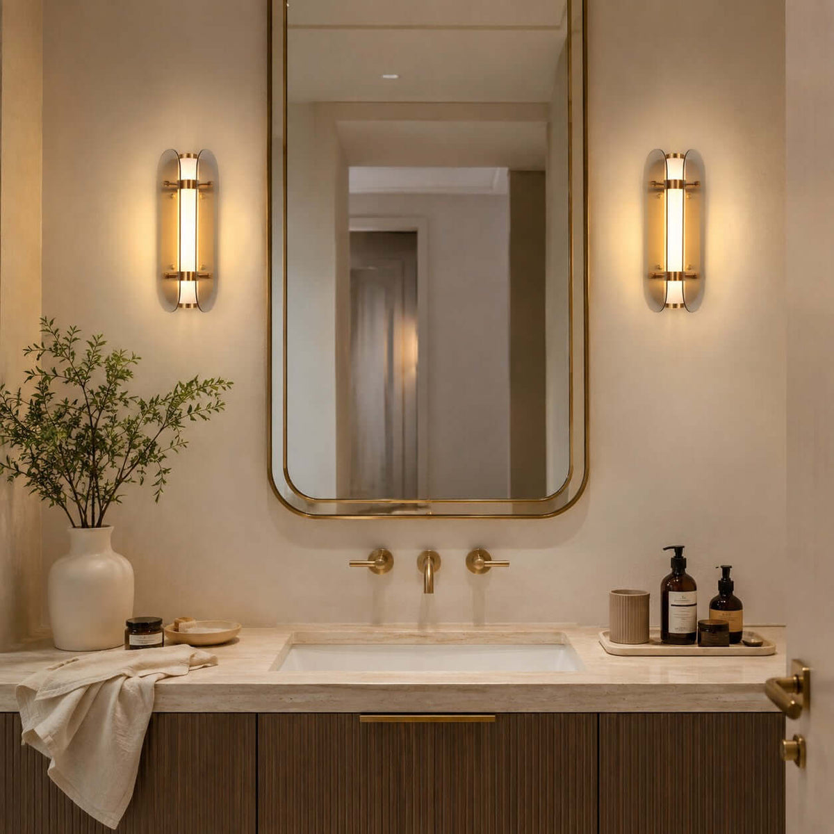 A pair of lit Clérachic wall lights flank a tall brass-framed mirror above a stone bathroom vanity with brass taps.