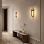 Clérachic wall lights lit on a panelled hallway wall, above a low stone bench and a vase of twigs.