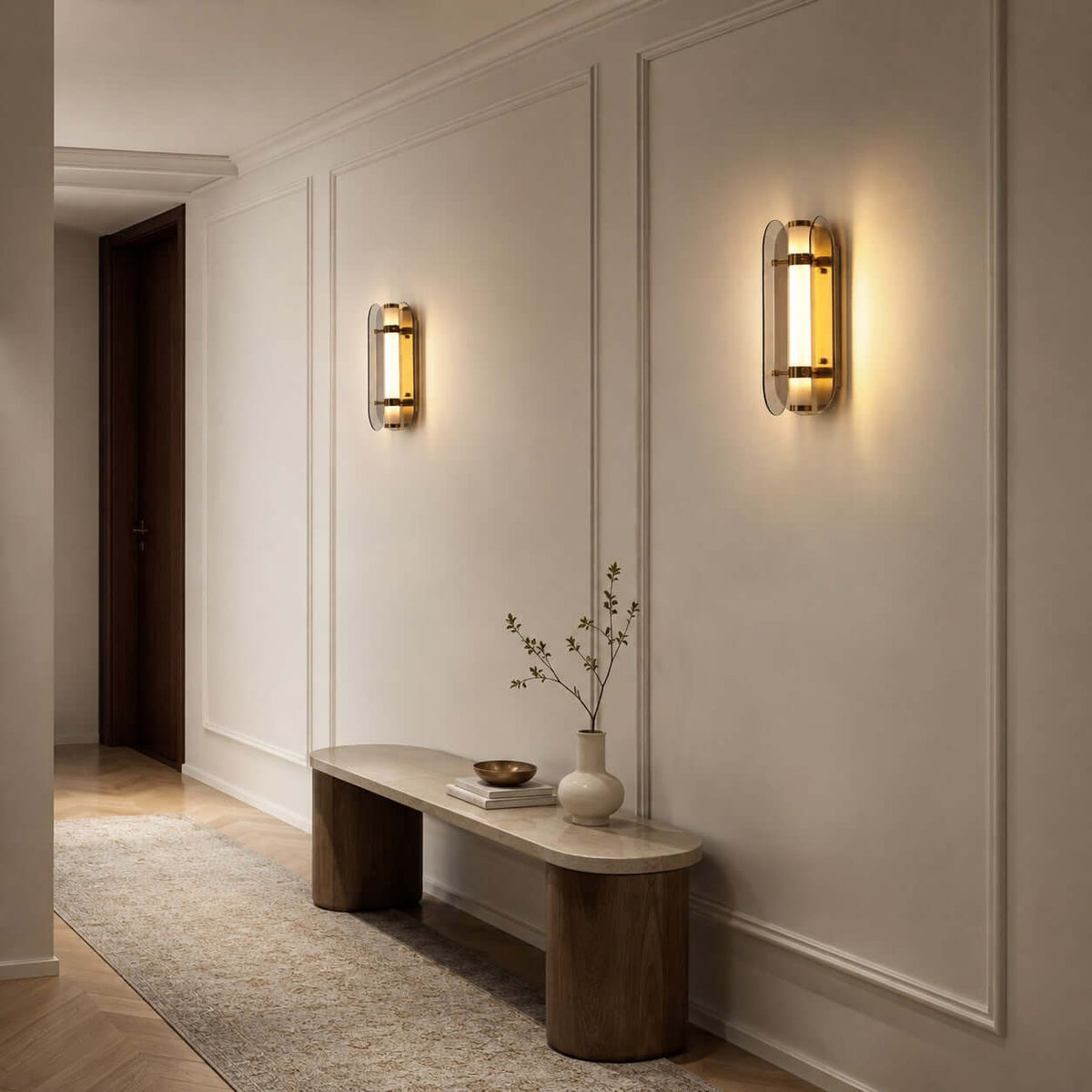 Clérachic wall lights lit on a panelled hallway wall, above a low stone bench and a vase of twigs.
