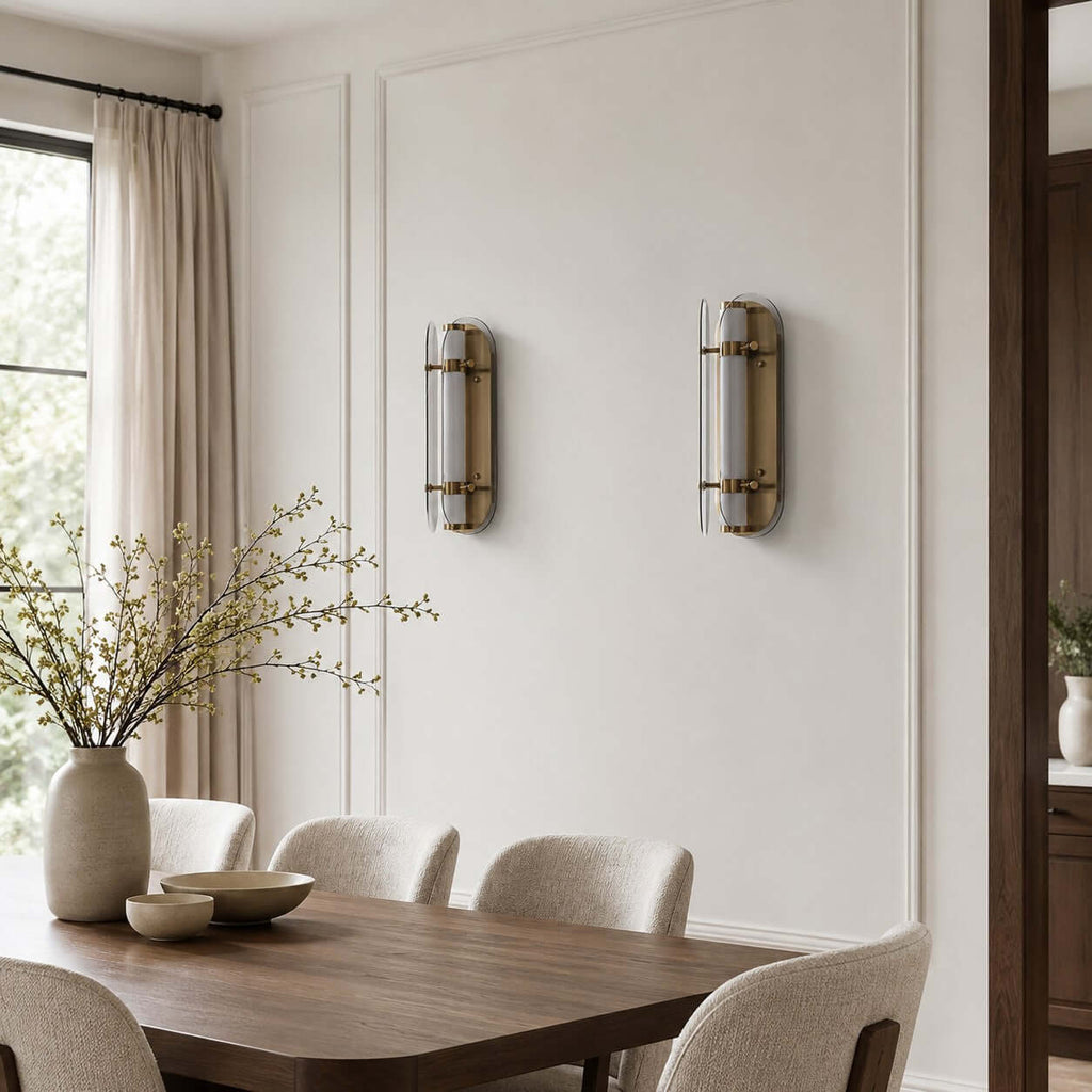 Two unlit Clérachic wall lights on a panelled wall above a wooden dining table, with boucle chairs and a vase of blossom.