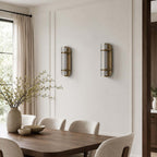 Two unlit Clérachic wall lights on a panelled wall above a wooden dining table, with boucle chairs and a vase of blossom.