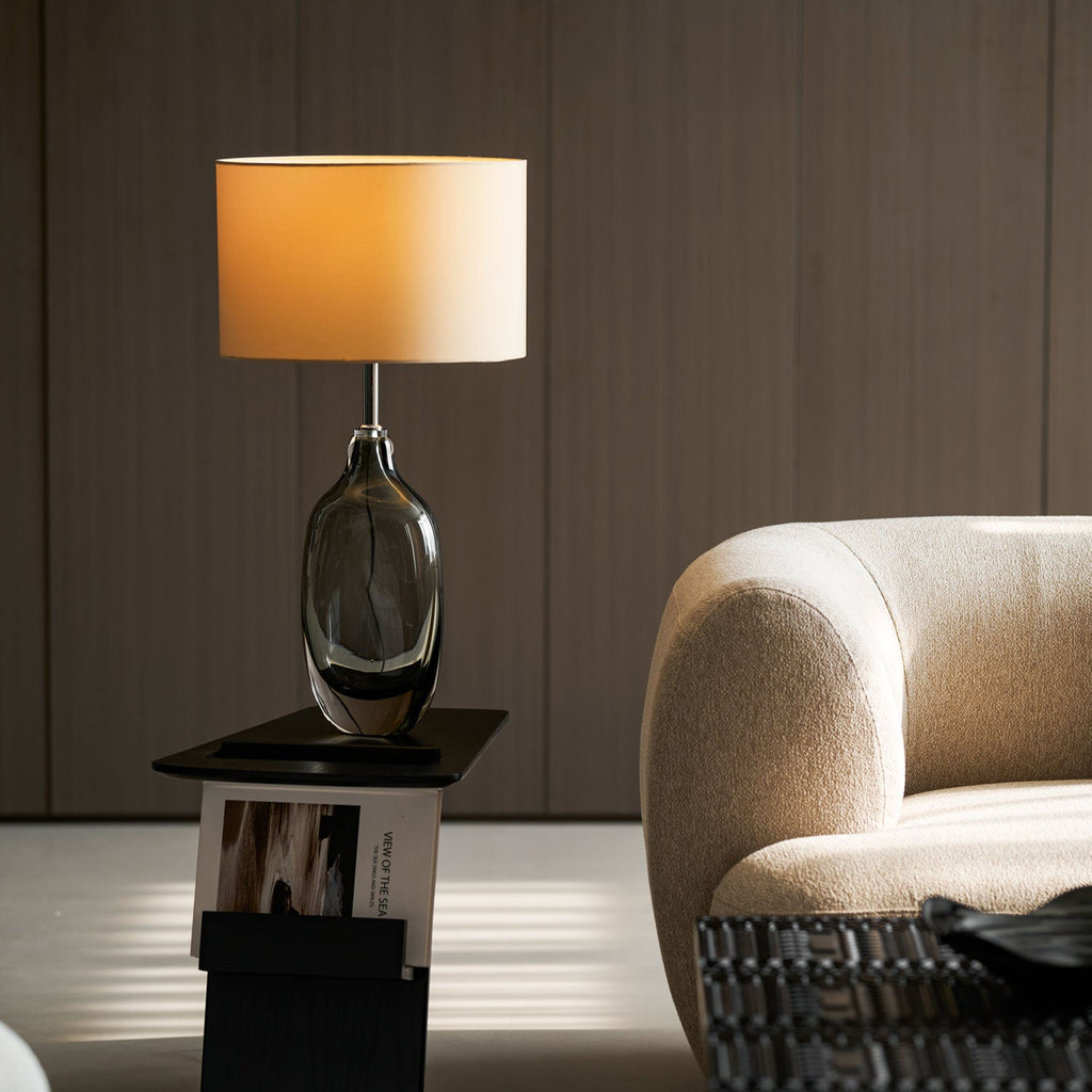 A lit Clérachic table lamp with a smoky glass base sits on a dark stand, with a boucle armchair and wood-slat wall behind.