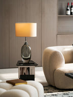 A Clérachic table lamp lit in a living room, with wine bottles on a shelf behind and a boucle sofa and pouffe in front.