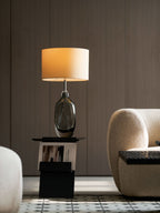 A Clérachic lamp with a smoky glass base and a lit drum shade stands before wood panelling, with a cream chair at its side.