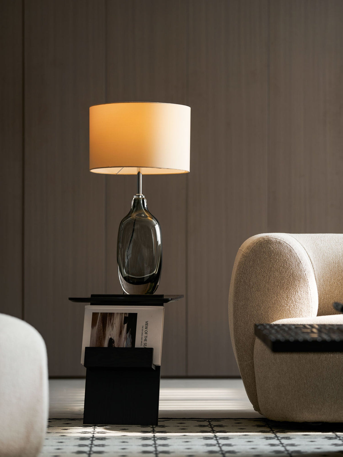 A Clérachic lamp with a smoky glass base and a lit drum shade stands before wood panelling, with a cream chair at its side.
