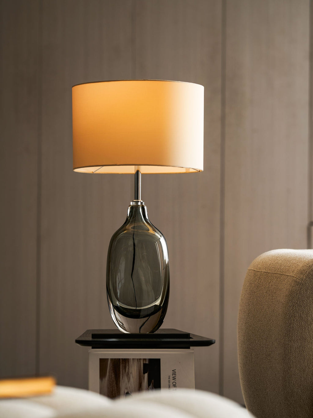 Close view of the lit drum shade and smoky glass body of this Clérachic table lamp, set against warm brown wall panelling.