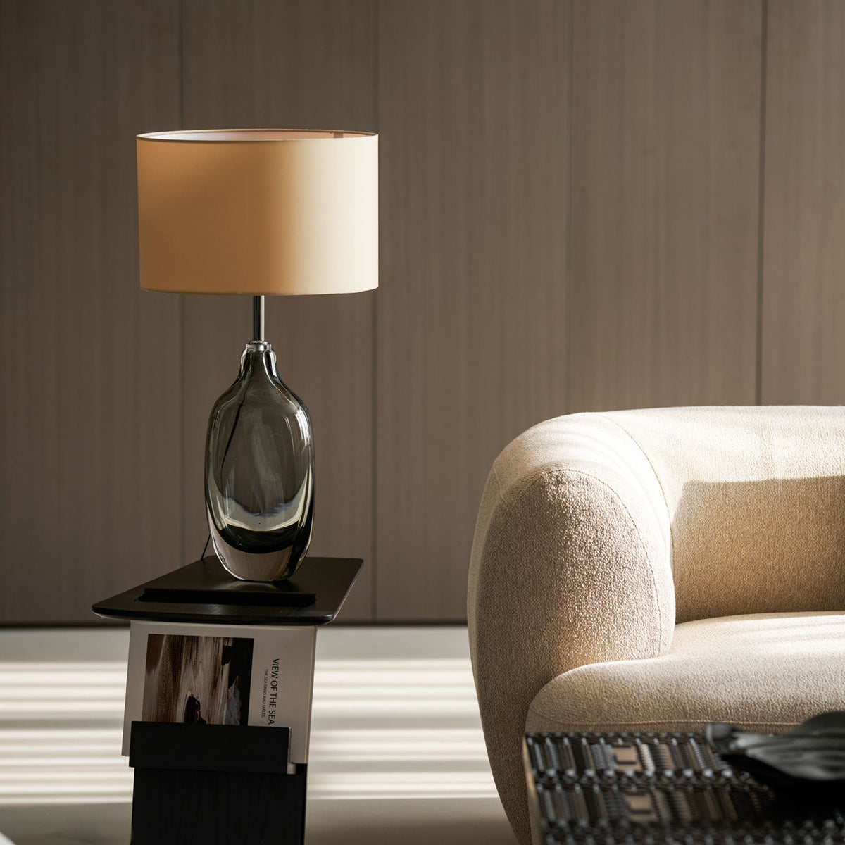 The Clérachic table lamp unlit on a dark side table, its cream shade catching daylight beside a rounded cream sofa.