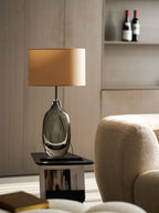 A Clérachic table lamp with a tan shade and smoky glass base on a black side table, with wine bottles on a shelf behind.