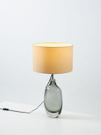 A Clérachic table lamp lit on a pale surface, with a glowing cream shade over the smoky grey glass base and an in-line switch on the cord.
