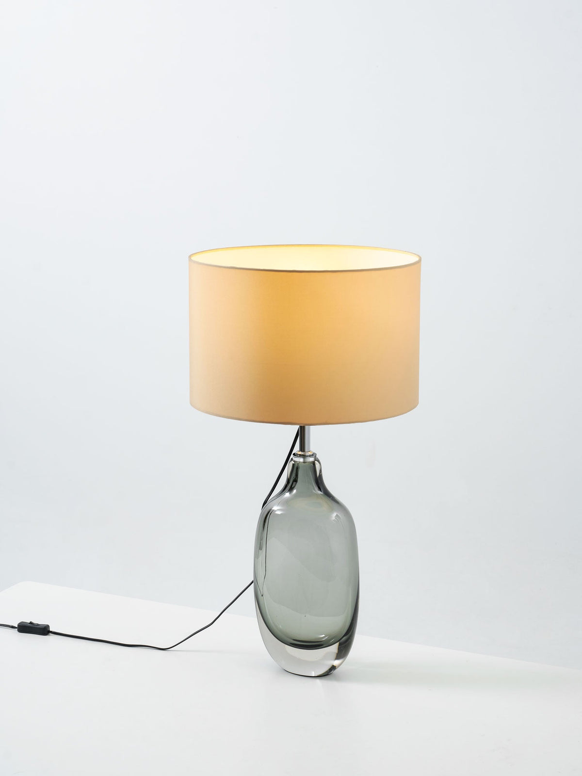 A Clérachic table lamp lit on a pale surface, with a glowing cream shade over the smoky grey glass base and an in-line switch on the cord.