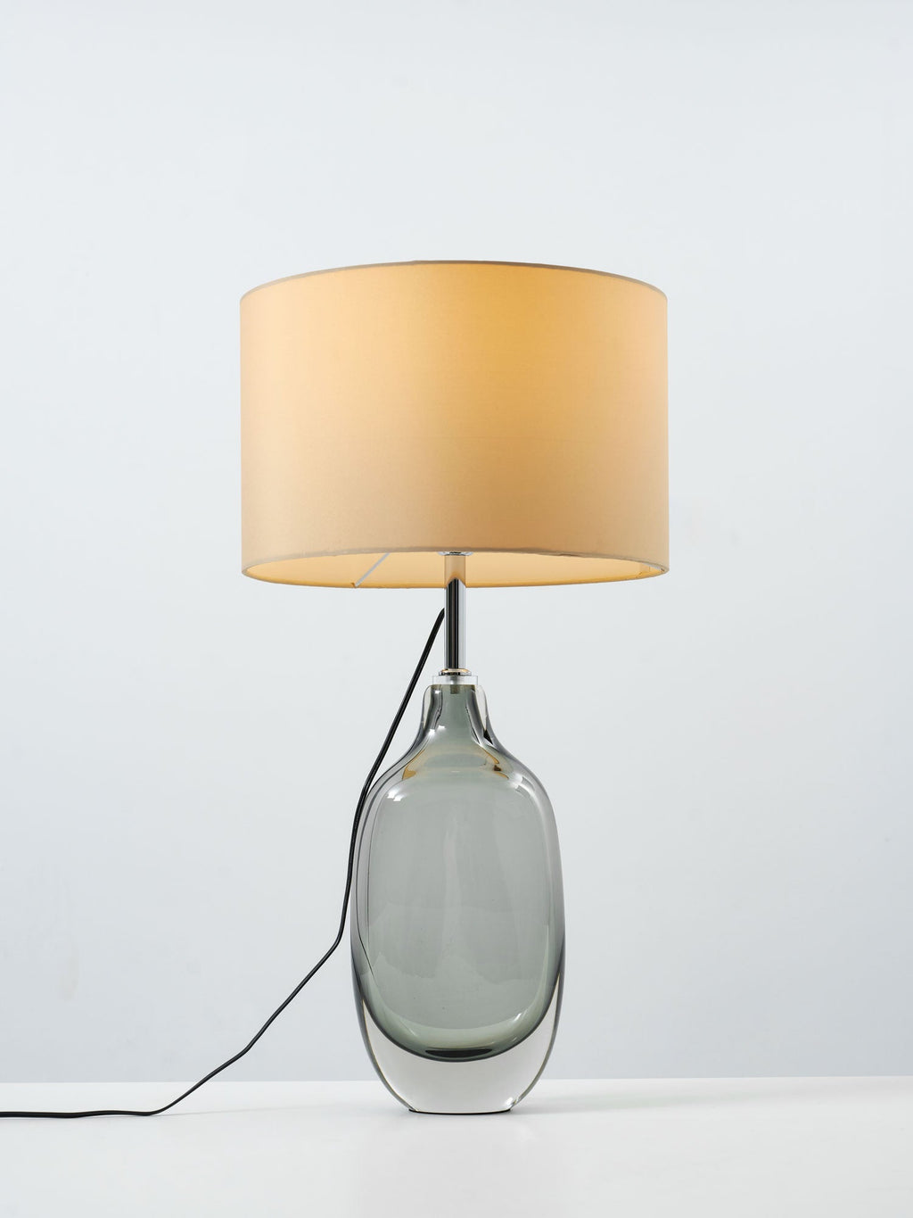 The Clérachic table lamp lit on a pale grey backdrop, showing the warm glow inside its wide cream drum shade.