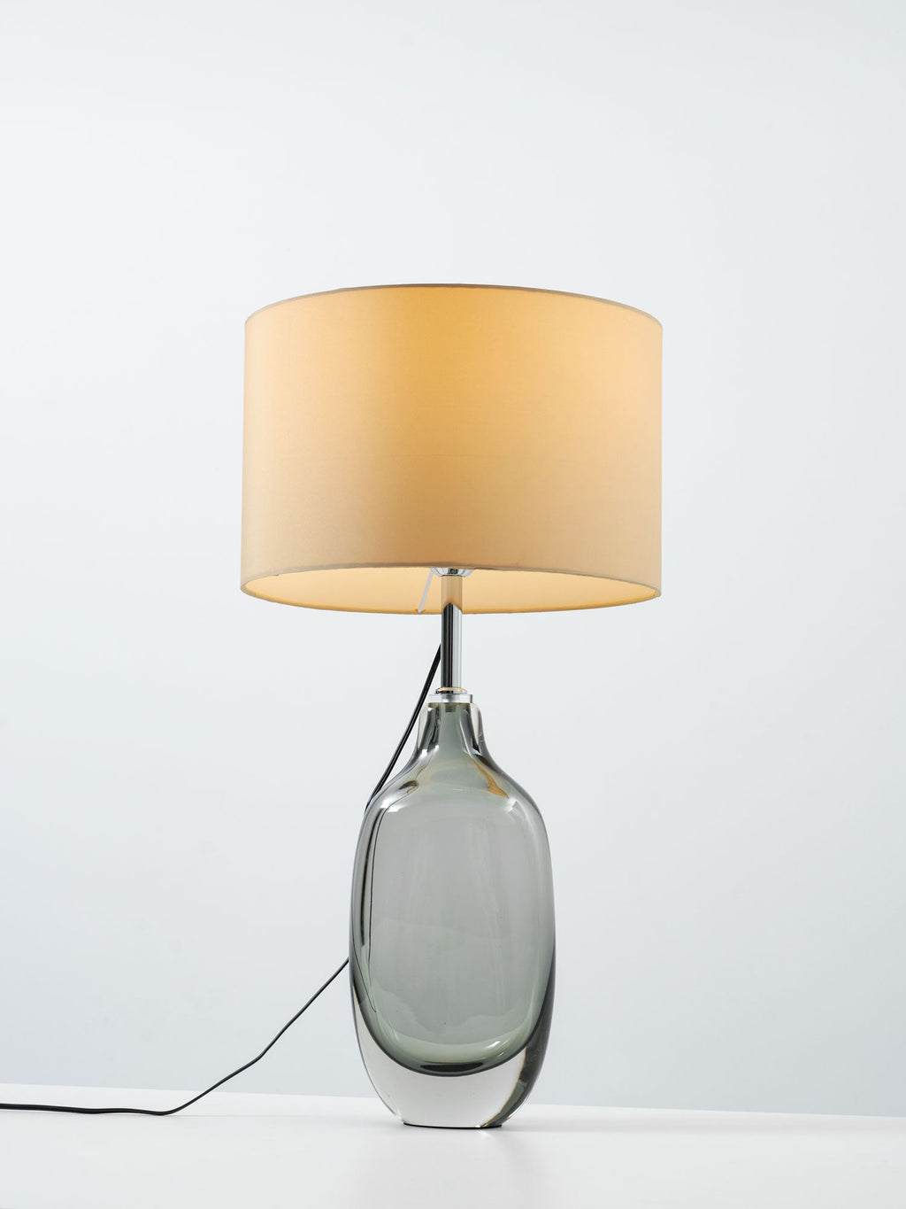 A taller, closer view of the lit Clérachic table lamp, with its cream drum shade over a thick-footed smoky glass base.