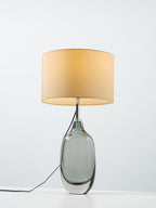 A taller, closer view of the lit Clérachic table lamp, with its cream drum shade over a thick-footed smoky glass base.