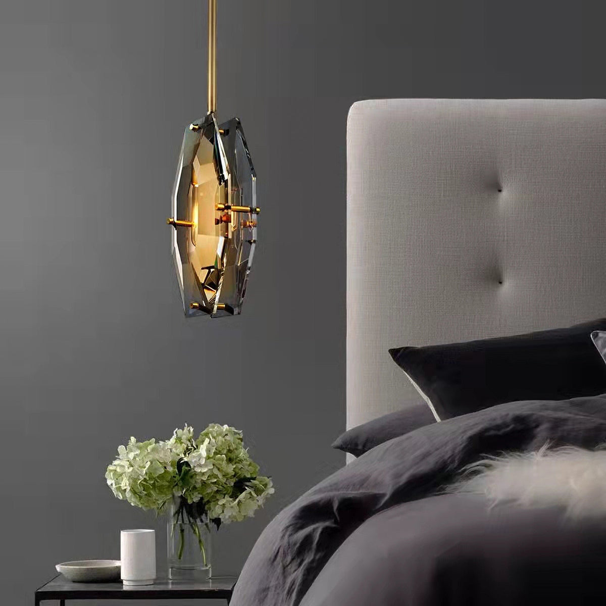 Smoky grey faceted pendant from Clérachic hanging beside a bed with a grey tufted headboard, dark bedding and a vase of white hydrangeas.