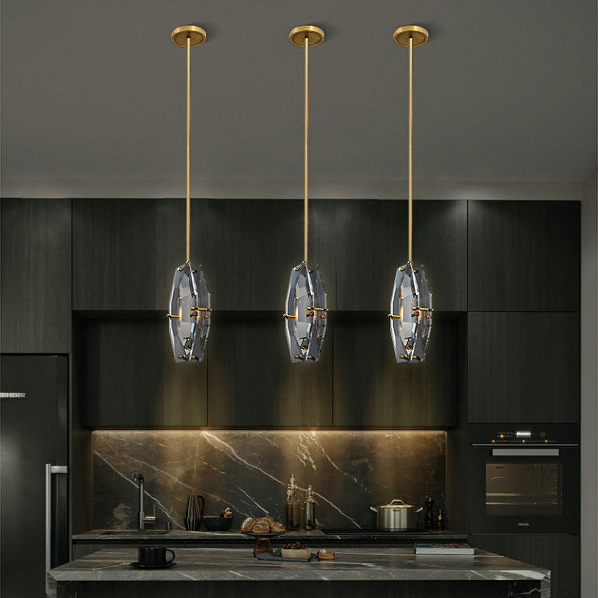 Three Clérachic crystal pendants hanging at one height on brass rods above a dark kitchen island, with a marble splashback behind.