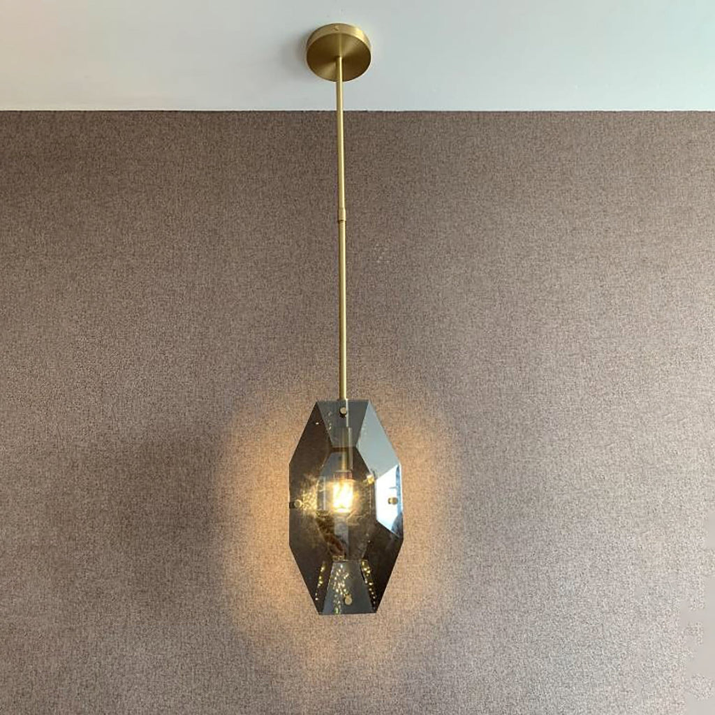 Lit Clérachic pendant on a brass rod and round ceiling plate in front of a textured brown wall, glowing warmly through smoky panels.