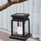 Clérachic black solar lantern with a clear glass body, candle bulb and metal handle, hanging from a tree branch by day.