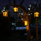 Four Clérachic black solar lanterns hung from tree branches and a post by their handles, glowing amber after dark.