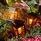 Clérachic black solar lanterns standing among pink flowers and green foliage, their candle bulbs glowing amber at dusk.