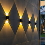 Four Clérachic black solar wall lamps in a diagonal run on a concrete wall, each casting warm triangles of light up and down at dusk.