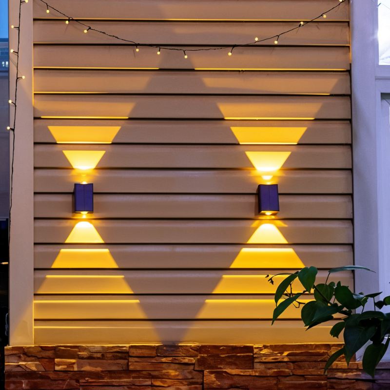 Clérachic pair of black solar wall lamps on pale siding under string lights, with warm triangles of light above and below.