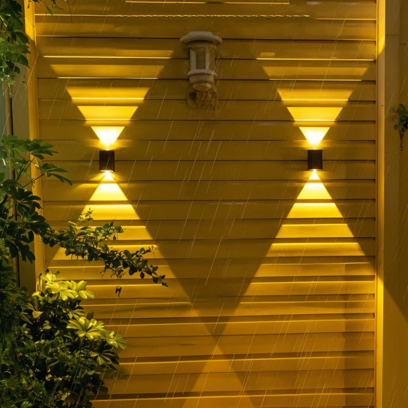Two small Clérachic black solar wall lamps on slatted wood in light rain, lighting the boards in warm triangles beside a porch light.