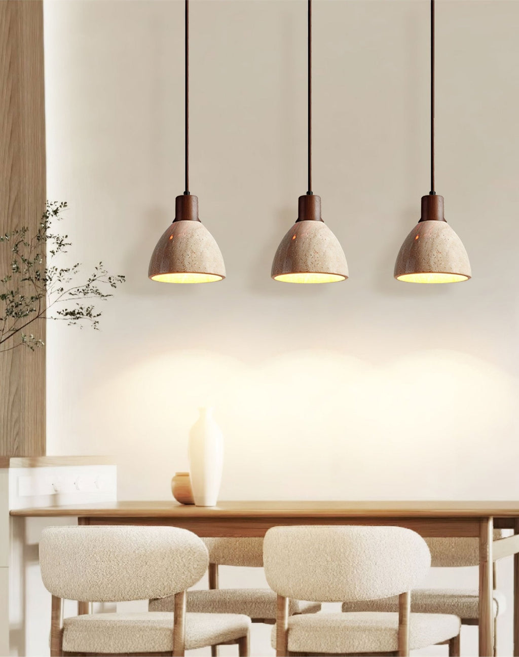 Three Clérachic travertine pendants in a row above a wooden dining table with cream bouclé chairs, each dome lit from below.