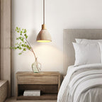 A Clérachic travertine pendant with a pale wood collar above a wooden bedside table, beside a grey headboard and a glass vase with a branch.