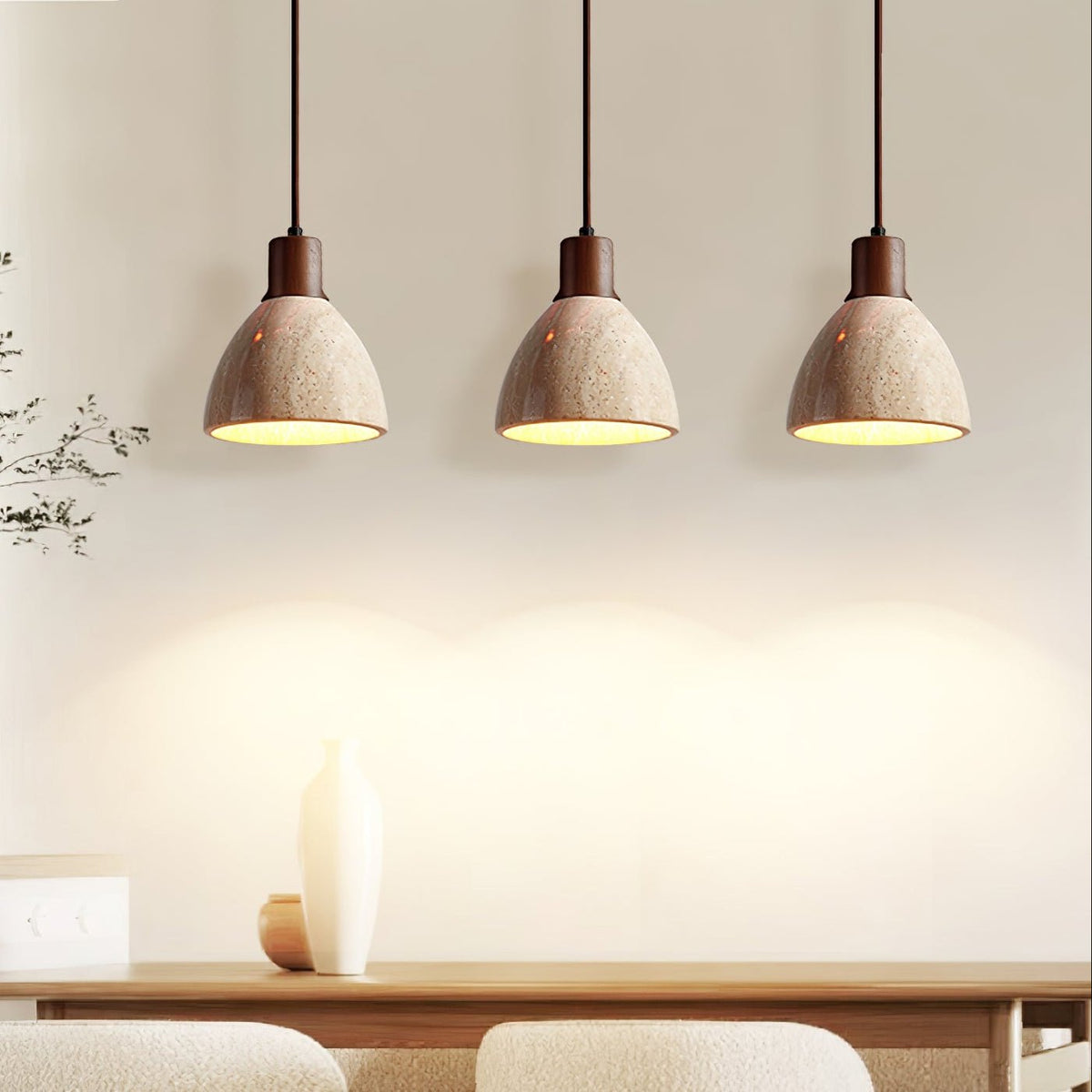 Three Clérachic pendants with dark walnut collars above a dining table, their travertine domes lit and glowing against a cream wall.