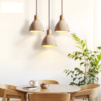 Three Clérachic pendants with pale wood collars hanging above a round oak table with a cup, with a leafy plant at the right edge.