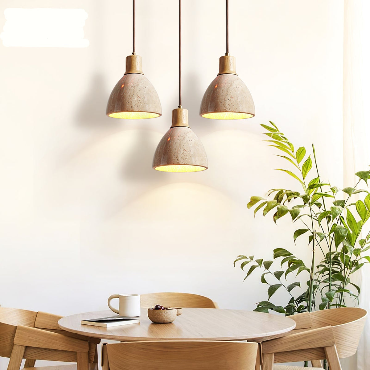 Three Clérachic pendants with pale wood collars hanging above a round oak table with a cup, with a leafy plant at the right edge.