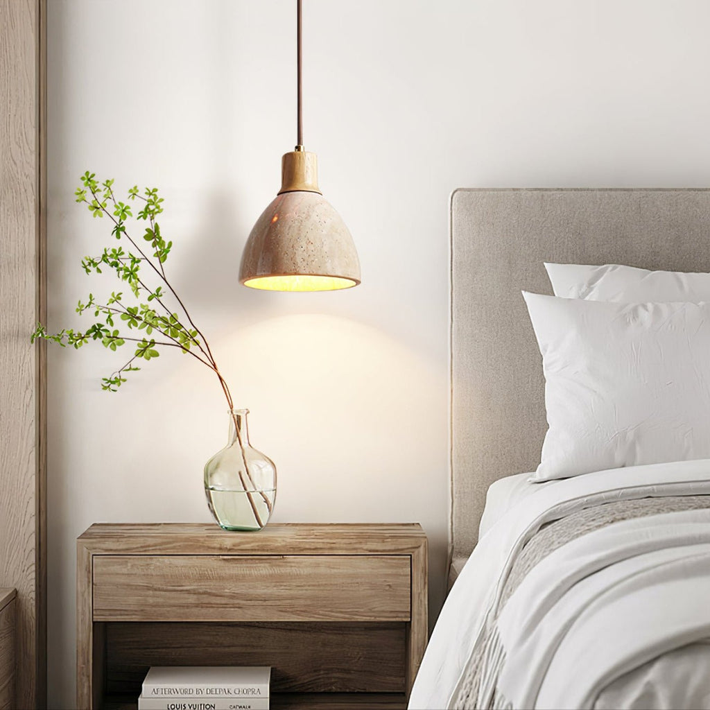 A Clérachic pendant with a pale wood collar above a bedside table with a glass vase and a branch, beside a bed with white pillows.