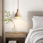 A Clérachic pendant with a pale wood collar above a bedside table with a glass vase and a branch, beside a bed with white pillows.
