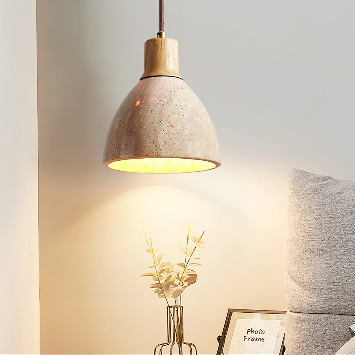 Low angle on a Clérachic travertine dome pendant lit warm, with a grey cushion, a gold wire vase of leaves and a photo frame beneath.