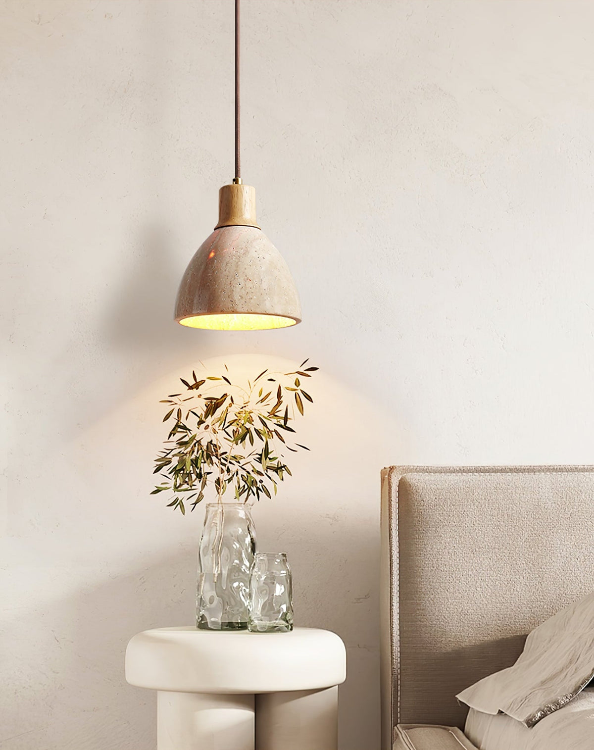 This Clérachic pendant with a pale wood collar glowing above a white stool and a glass vase of dried leaves, next to a taupe headboard.