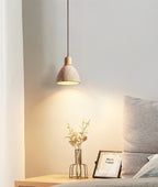A small Clérachic pendant glowing above a bedside shelf with a gold wire vase of leaves, a photo frame and a grey cushion.