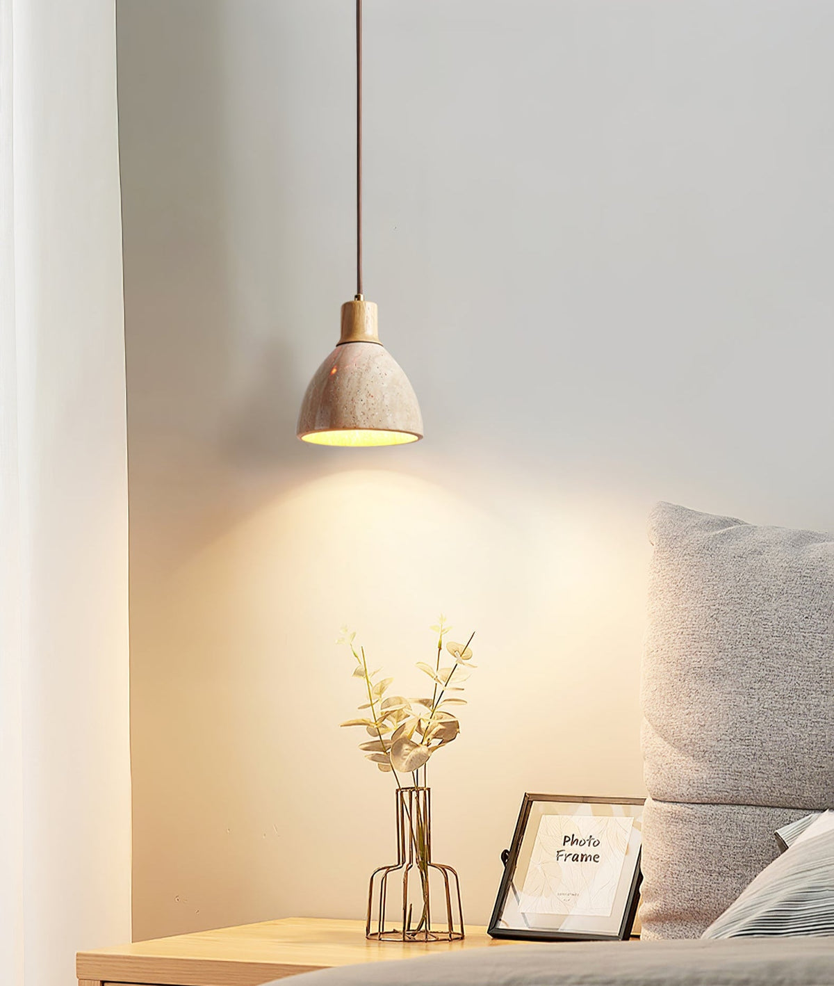 A small Clérachic pendant glowing above a bedside shelf with a gold wire vase of leaves, a photo frame and a grey cushion.