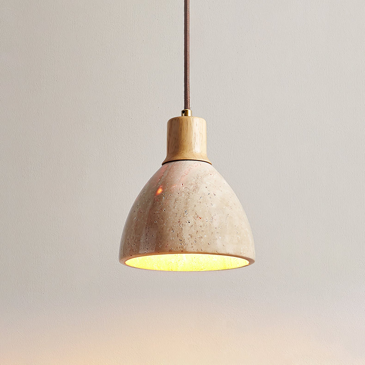 A Clérachic travertine pendant with a pale wood collar on a plain taupe wall, the underside of the dome glowing yellow-gold.