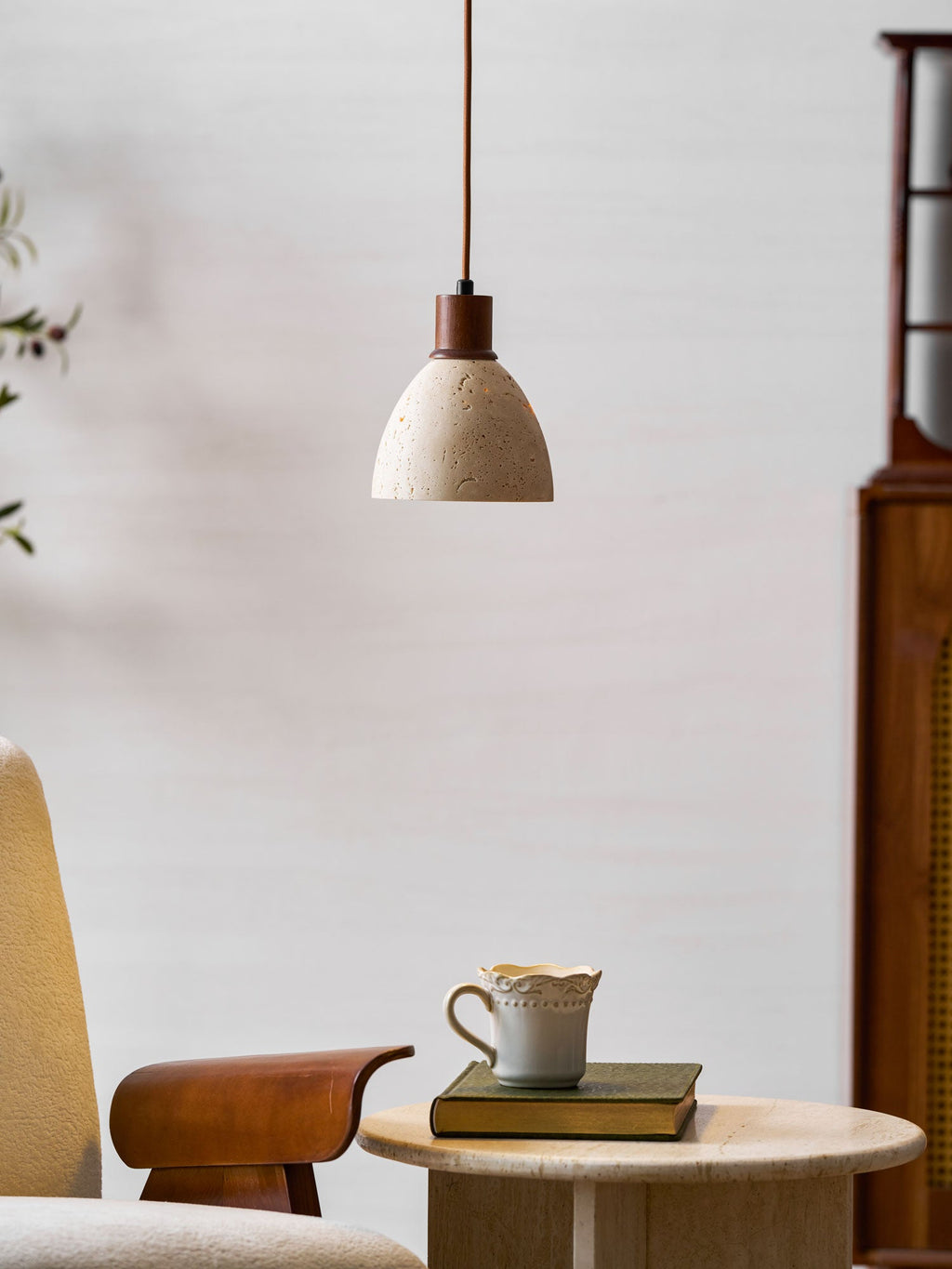 A Clérachic pendant with a dark walnut collar hanging above a round travertine side table with a cup and a green book.
