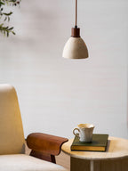 This Clérachic pendant with a walnut-toned collar above a cream armchair with a wooden armrest and a side table holding a book and a cup.