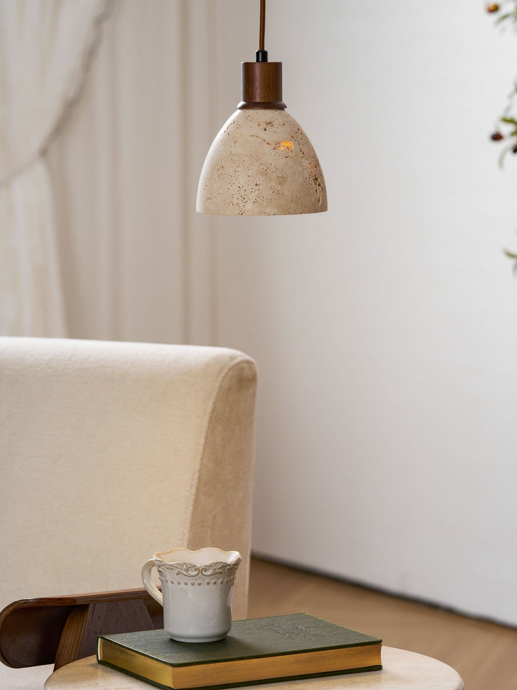 A Clérachic travertine dome pendant above a cream armchair and a travertine side table with a lace-edged cup and a book.