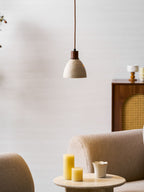 A Clérachic pendant hung above a beige armchair and a round side table with yellow candles, a small cup and a cane cabinet behind.