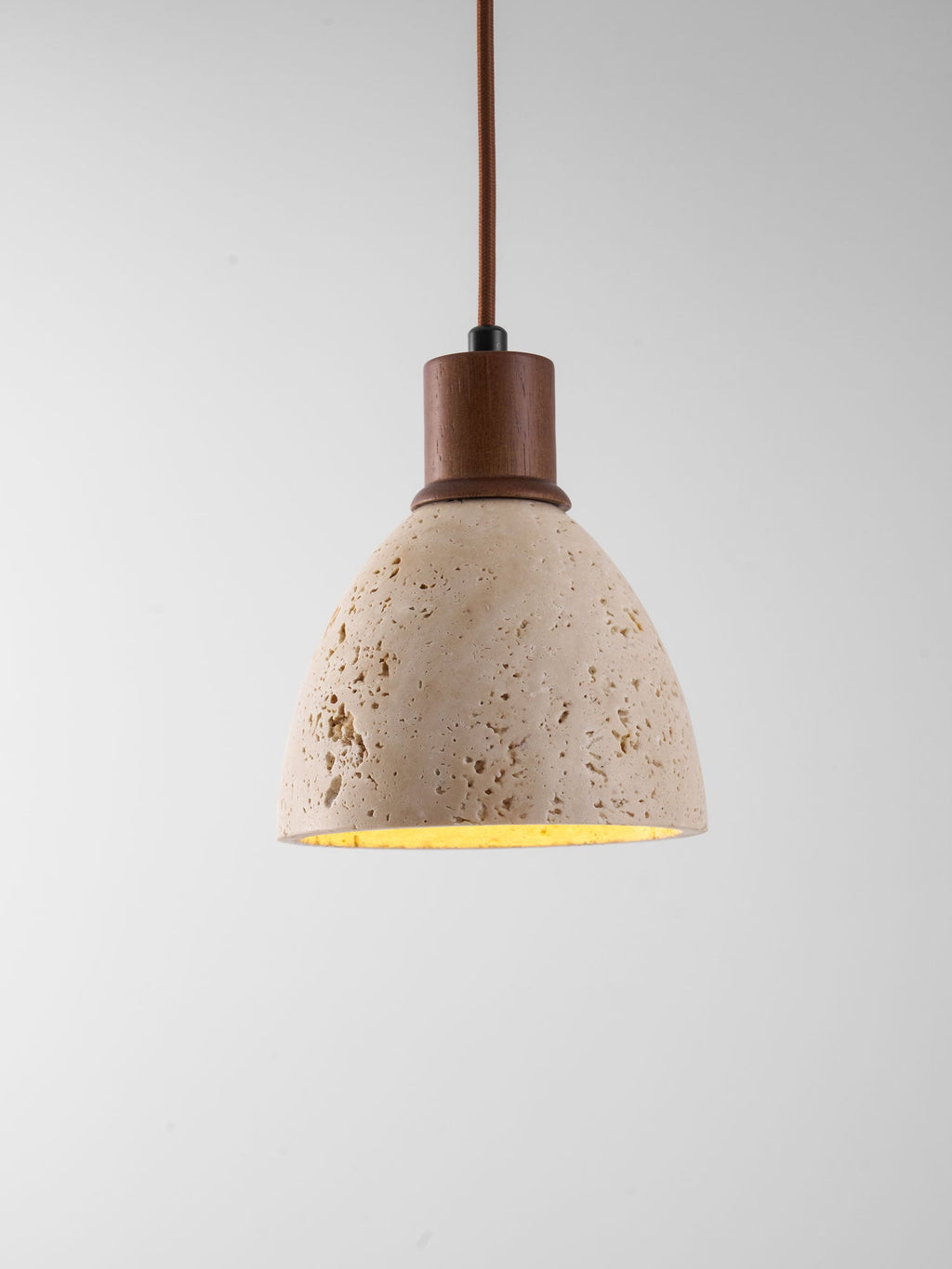 A Clérachic pendant on a grey backdrop, its pitted travertine dome lit from inside under a dark walnut collar and brown cord.