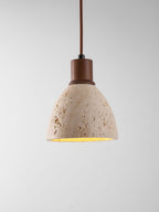 A Clérachic pendant on a grey backdrop, its pitted travertine dome lit from inside under a dark walnut collar and brown cord.