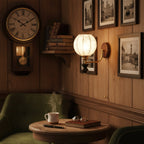Clérachic wall light with a round fabric shade glowing on a wood-panelled wall, beside a wall clock, framed prints and a green armchair.
