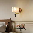 Clérachic wall light with a pale printed drum shade, set on a cream wall beside a wooden headboard and a vase of pink roses.