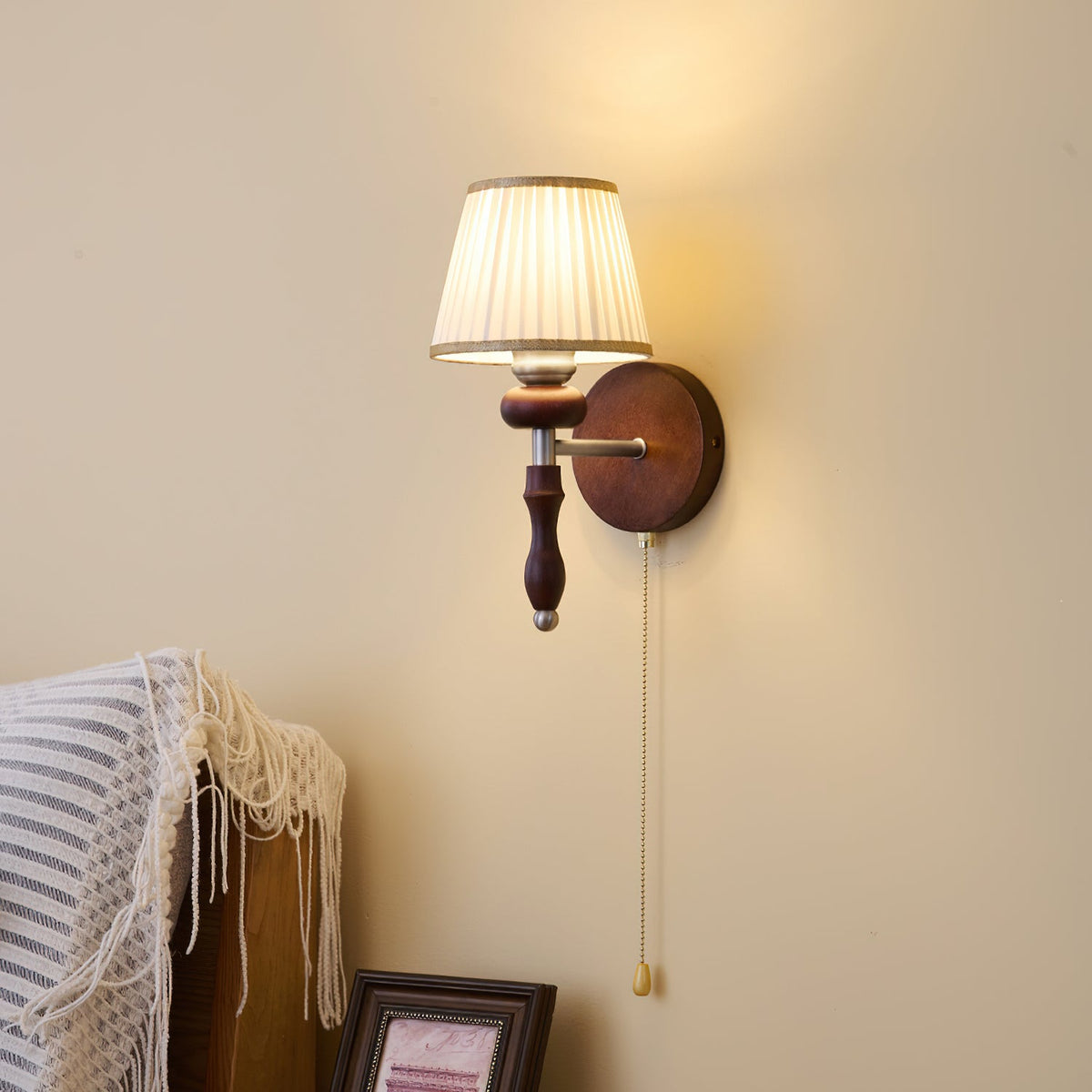 Clérachic wall light with a pleated white shade and a dark wood stem, lit against a cream wall beside a striped throw.